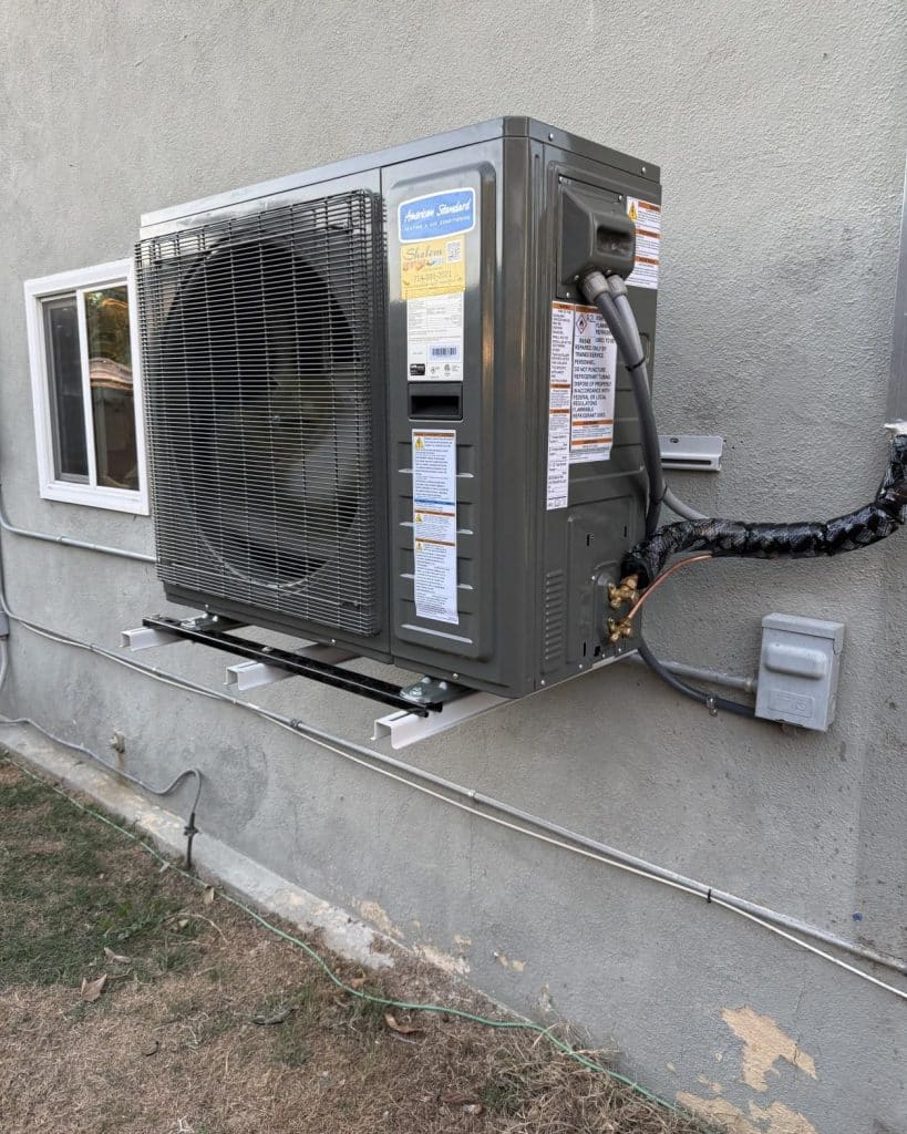 American Standard mini-split heat pump condenser mounted on wall bracket against gray stucco with refrigerant lines.