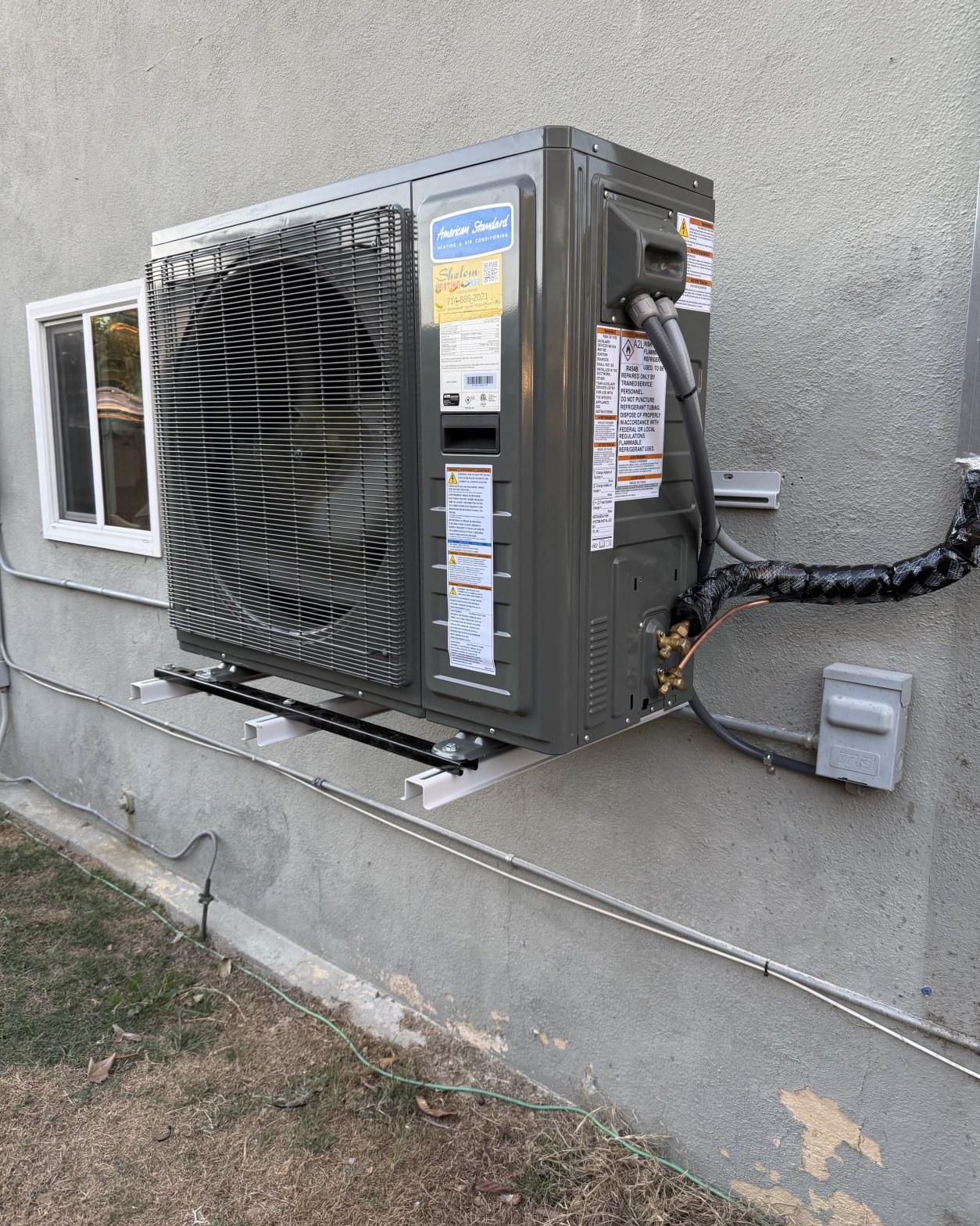 American Standard mini-split heat pump condenser mounted on wall bracket against gray stucco with refrigerant lines.